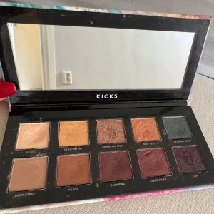 Festival Collection Eyeshadow Palette från Kicks - Säljer en färgglad ögonskuggspalett från Kicks Festival Collection med 10 olika nyanser, både matta och skimrande. Paletten har en stor spegel och nyanser i beige, guld, brons, grönt och vinrött. Perfekt för kreativa sminkningar.