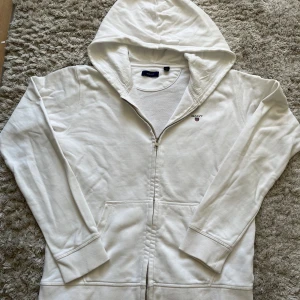 Gant hoodie - Säljer en klassisk vit hoodie från GANT med dragkedja framtill och broderad logga på bröstet. Tröjan har huva, långa ärmar och två fickor framtill. Perfekt för alla outfits och en avslappnad stil. Den är storlek 170 / 15 Years men skulle säga att den passar XS.