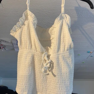 Vit croppad linnetopp med volang från SHEIN - Säljer en söt vit croppad linnetopp från SHEIN med volangdetaljer längs axelbanden och v-ringning. Toppen har knytning framtill och är tillverkad i ett lätt, strukturerat material. Perfekt för varma dagar!