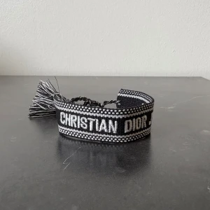 Svart armband från Christian Dior - Säljer ett snyggt armband från Christian Dior i svart och vitt med broderad logga och tofsdetaljer. Armbandet har justerbar knytning och är en perfekt accessoar för att lyfta din outfit.
