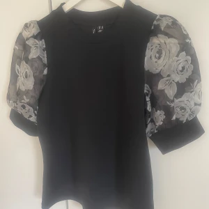 Svart topp med blommiga puffärmar från Vero Moda - Säljer en svart topp från Vero Moda med genomskinliga puffärmar i blommigt mönster. Toppen har rund halsringning och en tight passform. Perfekt för dig som vill ha något unikt och stilrent!