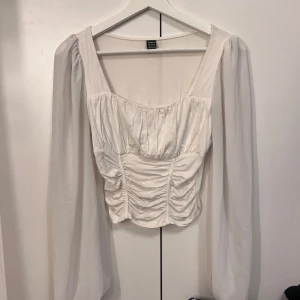 Vit långärmad topp med rynkade detaljer  - Vit långärmad blus med rynkade detaljer framtill, fyrkantig ringning och chiffongärmar. Toppen har en figurnära passform, använd en gång.