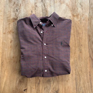 Ralph Lauren – Rutig skjorta i vinröd/grön flanell - Klassisk button-down-skjorta i mjuk flanell med mörkgrön broderad logga. Perfekt för lager-på-lager eller för sig själv – tidlös stil med preppy känsla.  Storlek: L (normal i storlek) Färg: Vinröd, grön och gul rutig med mörkgrön logga Material: 100% bomull (flanell) Skick: Mycket fint skick – inga hål eller fläckar Stil: Preppy / Casual / Vintage Passform: Klassisk passform med button-down-krage och rundad fåll Nypris: ca 1 400 kr Pris: 300 kr Frakt: Skickas snabbt & spårbart
