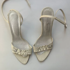 Vita  sandaler med klack  - Säljer ett par vita sandaler från Pablo Santoni med smal klack och öppen tå. Skorna har ett rynkat band framtill med spänndetalj i silver och justerbar rem runt vristen. Storlek 39.