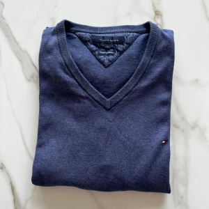 Blå v-ringad tröja från Tommy Hilfiger - Säljer en klassisk blå v-ringad tröja från Tommy Hilfiger. Tröjan är tillverkad i en mjuk bomulls- och kashmirblandning och har långa ärmar samt en liten broderad logga på bröstet. Perfekt för en stilren och avslappnad look.