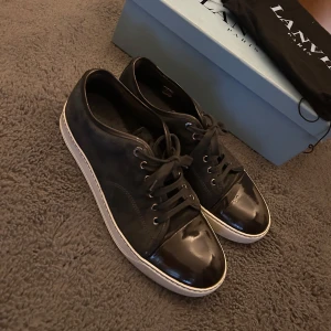 Lanvin skor  - Säljer ett par svarta sneakers från Lanvin med snygg lackad tå och vit sula. Skorna har snörning och en stilren design i mocka och skinn. Perfekta för dig som vill ha en lyxig och clean look. Ovanlig colorway elephant grey, storlek 43 men sitter mer som 44-45. Hör av dig vid minsta lilla fundering!