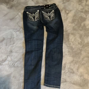 Mörkblå jeansbyxor från Miss Me med broderade bakfickor - Säljer ett par mörkblå jeans från Miss Me med snygga kontrastsömmar och detaljerade broderier på bakfickorna. Byxorna har låg midja och raka ben. Perfekta för dig som gillar statement-jeans!