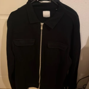 Svart overshirt med dragkedja - Säljer en svart overshirt med klassisk krage och två bröstfickor. Jackan har en rak passform och stängs med en silverfärgad dragkedja framtill. Perfekt för lager-på-lager-stil. Sitter som L