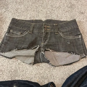 Lågmidjade jeansshorts  - Säljer ett par svarta jeansshorts från Vero Moda med råa, fransiga kanter och dubbla knappar framtill. Shortsen har klassiska fickor både fram och bak samt vita kontrastsömmar för en cool look.