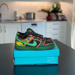 Nike Dunk Low med färgglada detaljer - Säljer ett par Nike Dunk Low med svart bas, gröna swoosh och sulor samt bruna och orange detaljer. Skorna har unika grafiska mönster i gult, rosa och blått på hälen. Perfekta för dig som vill sticka ut med dina sneakers!