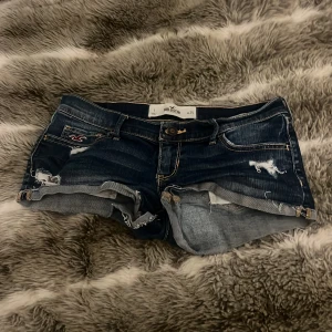 Mörkblå jeansshorts från Hollister - Snygga mörkblå jeansshorts från Hollister med slitna detaljer och råa kanter. Klassisk femficksmodell med Hollister-logga på ena fickan och bakfickor med dekorativa sömmar. Perfekta för sommaren! Lågmidjade också!!!!!
