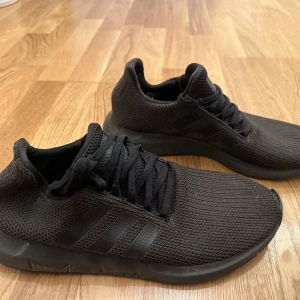 Svarta sneakers från Adidas - Säljer ett par helsvarta Adidas sneakers med stickad ovandel och klassiska tre ränder på sidan. Skorna har snörning och platt sula, perfekt för en sportig och stilren look.
