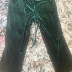 Gröna sammetsbyxor  - Mörkgröna sammetsbyxor  med resår i midjan och knytband. Byxorna har en loose passform och är perfekta för en avslappnad stil. Materialet är mjukt och glansigt, vilket ger en lyxig känsla.