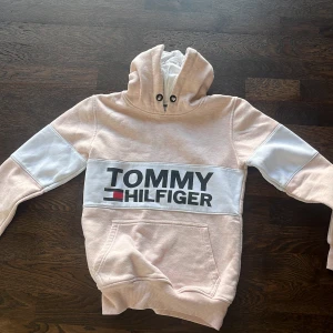 Ljusrosa hoodie från Tommy Hilfiger - Säljer en ljusrosa hoodie från Tommy Hilfiger med vit panel och stor logga framtill. Tröjan har huva med snörning, magficka och vita detaljer på ärmarna. Perfekt för en avslappnad och snygg stil.