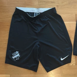 Svarta fotbollsshorts från Nike AIK - Svarta fotbollsshorts från Nike med AIK-logga på ena benet och vit Nike-swoosh på det andra. Tillverkade i Dri-Fit-material som andas och har resår i midjan. Perfekta för fotbollsträning eller match.