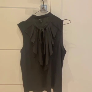 Svart ärmlös blus med volang och knyt - Säljer en svart ärmlös blus med volangdetaljer framtill och knyt vid halsen. Perfekt för dig som gillar en elegant och stilren look. Blusen har en mjuk och lätt känsla.