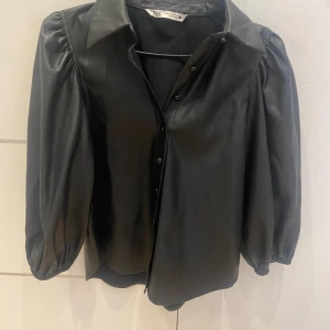 Svart skjorta i skinnimitation från Zara - Säljer en svart skjorta i skinnimitation från Zara med puffiga långärmar och klassisk krage. Skjortan har knappar framtill och en snygg, lite glansig finish. Perfekt för dig som vill sticka ut med en trendig look.