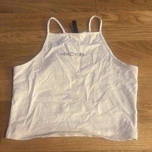 Vit croppad linnetopp från Divided - Säljer en vit croppad linnetopp från Divided med tunna axelband och trycket 'New York' framtill. Perfekt för varma dagar och enkel att matcha med jeans eller kjol.