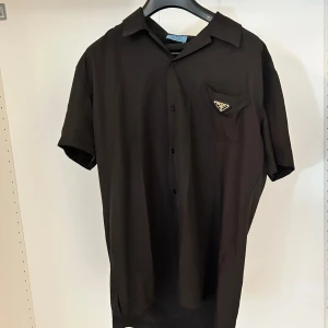Prada kortärmad skjorta  - Snygg svart skjorta från Prada med korta ärmar och klassisk krage. Skjortan har en bröstficka med Pradas ikoniska metallogo. Perfekt för dig som gillar stilrena och exklusiva plagg.