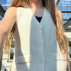 Beige stickad väst med knappar - Säljer en beige stickad väst med v-ringning och fyra stora knappar framtill. Västen har två dekorativa fickor och är ribbad i materialet. Perfekt att styla över en t-shirt eller skjorta för en trendig lager-på-lager-look.