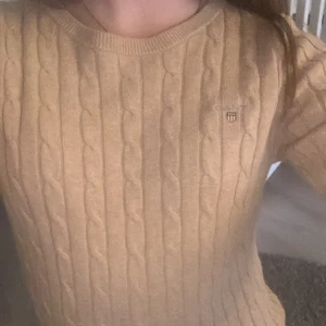 Beige kabelstickad tröja från Gant - Säljer en klassisk beige kabelstickad tröja från Gant med rund halsringning och diskret logga på bröstet. Tröjan har lång ärm och är perfekt för lager-på-lager-stil. Supermjuk och stilren för en snygg vardagslook.