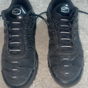 Nike Air Max Plus Tn helsvarta sneakers - Säljer ett par helsvarta Nike Air Max Plus Tn sneakers med klassisk Tn-detalj på hälen och vågformade linjer på ovandelen. Skorna har snörning, rund tå och platt sula. Perfekta för dig som gillar en sportig och stilren look.