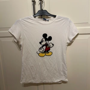 Vit t-shirt med Mickey Mouse från Disney - Vit t-shirt från Disney med ett stort motiv av Mickey Mouse framtill, dekorerad med pärlor i svart, vitt, rött och gult. Klassisk rund halsringning och korta ärmar. I strl XL (är liten i strl)