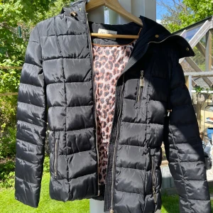 Svart pufferjacka med dragkedjor - Svart pufferjacka från Roco Baroco med quiltad design och flera dragkedjor framtill. Jackan har hög krage och långa ärmar, perfekt för kyligare dagar. Snygg och stilren modell som passar till mycket.