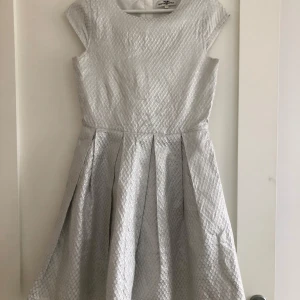 Ljus silverfärgad klänning med struktur - Säljer en ljus silverfärgad klänning med korta ärmar och markerad midja. Klänningen har ett strukturerat tyg med diskret mönster och utställd kjol som ger en fin siluett. Perfekt för speciella tillfällen!