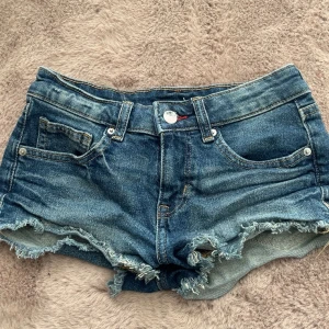 Blå låg midjade jeansshorts - Säljer ett par blå lågmidjade jeansshorts från H&M i storlek 34. Shortsen har låg midja, klassisk femficksdesign och råa fransade benslut för en avslappnad look. Perfekta för varma dagar och enkel att matcha med olika toppar.