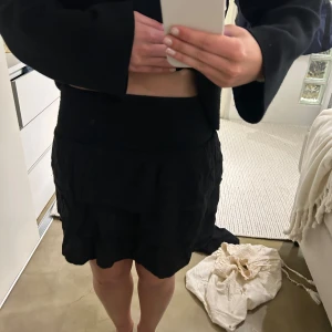 Svart kortkjol med volang - Säljer en svart kortkjol med volangdetalj. Kjolen har en lös passform och är perfekt för dig som gillar en enkel men ändå trendig stil. Passar till många olika outfits och är lätt att matcha.