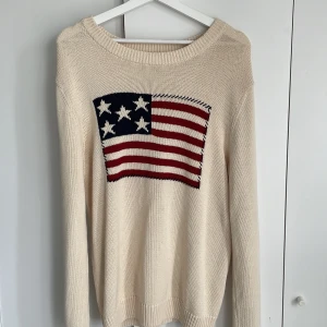 Beige stickad tröja med amerikansk flagga flagga från Selected Homme - Beige stickad tröja från Selected Homme med ett stort motiv av den amerikanska flaggan på bröstet. Tröjan har rund halsringning och långa ärmar. Perfekt för dig som gillar klassiska och stilrena plagg med en twist.