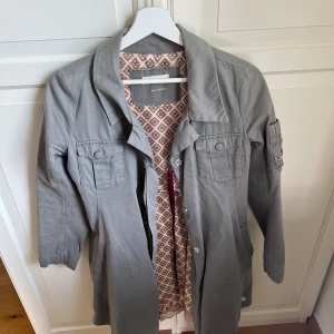 Grå kappa med broderad detalj - Säljer en grå kappa från Odd Molly med broderad patch på ena ärmen och fina fickdetaljer framtill. Kappan har knappar, krage och ett mönstrat innerfoder. Perfekt för dig som vill ha något unikt och stilrent.