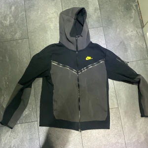 Svart och grå zip-hoodie från Nike. Storlek S - Säljer en svart och grå hoodie från Nike med hel dragkedja och huva. Jackan har en gul Nike-logga på bröstet och detaljer i form av svarta ränder över bröstet. Ficka med dragkedja på ärmen och sportig look.