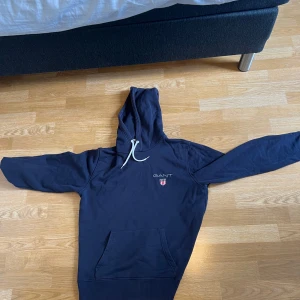 Mörkblå hoodie från GANT - Säljer en mörkblå hoodie från GANT med vit dragsko i huvan och klassisk logga broderad på bröstet. Tröjan har känguruficka framtill och långa ärmar. Perfekt för en avslappnad stil.