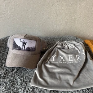 Grå keps från Lyxery by Sweden - Snygg beige keps från Lyxery by Sweden med mesh på sidorna och baksidan. Framsidan har ett svartvitt tryck av en staty och stadsmiljö. Kepsen har broderad logotyp och detaljer i metall samt justerbar passform. Original pris 899 från jeansbolaget mitt pris 399kr pris kan diskuteras 