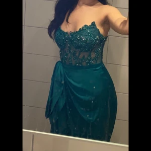 Grön långklänning med spets och paljetter - Säljer en elegant grön långklänning med v-ringad överdel i spets och paljetter. Klänningen är ärmlös och har en figurnära passform upptill med draperad kjol. Perfekt för bal eller fest.