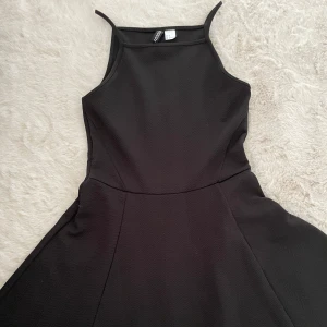 Svart klänning med smala axelband - Säljer en svart klänning med smala axelband och figurnära överdel. Klänningen har en enkel och stilren design med utsvängd kjol och hög halsringning. Perfekt för dig som gillar minimalistisk stil.