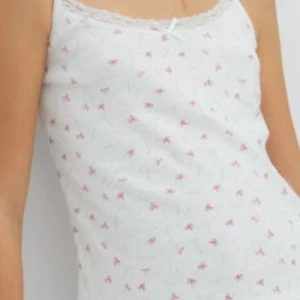 Vit linnetopp med små rosa rosetter och spetskant - Supersöt vit linnetopp med tunna axelband och små rosa rosetter som mönster. Toppen har en fin spetskant vid urringningen och en liten rosett framtill. Original pris 100kr💞