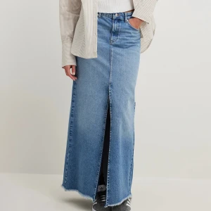 Ljusblå lång jeanskjol - Säljer en ljusblå lång jeanskjol med hög midja och rak passform. Kjolen har en snygg slits framtill och klassiska jeansdetaljer som fickor och bälteshällor. Perfekt att styla med sneakers eller boots. Kjolen är oanvänd och har prislappen kvar. 