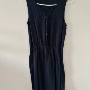 Marinblå ärmlös klänning med knyt - Säljer en stilren marinblå klänning med knyt framtill och resår i midjan. Klänningen är ärmlös och har en enkel, rak passform som passar perfekt till sommaren. Materialet känns mjukt och lätt, vilket gör den skön att bära varma dagar.