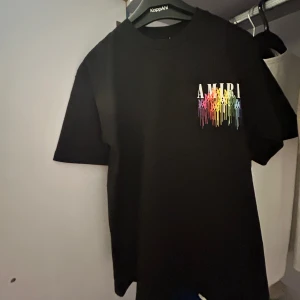 Svart t-shirt från Amiri med färgglatt tryck - Säljer en svart t-shirt från Amiri med färgglatt regnbågstryck och logga både fram och bak. T-shirten har normal passform och är kortärmad. Perfekt för dig som gillar streetwear och vill sticka ut med snygga detaljer.