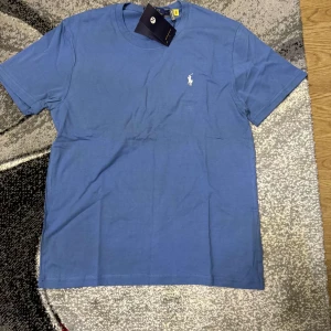 Blå t-shirt från Polo Ralph Lauren - Säljer en klassisk blå t-shirt från Polo Ralph Lauren med liten broderad logga på bröstet. T-shirten har rund halsringning och är tillverkad i mjuk bomull. Perfekt för en clean och stilren look.