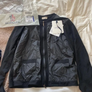 Svart cardigan jacka från Moncler - Svart cardigan jacka från Moncler med dragkedja framtill och två fickor. Jackan har långärmade ärmar och är tillverkad i en kombination av bomull och syntetmaterial. Använd vid enstaka tillfällen så i gott skick, digitalt kvitto finns