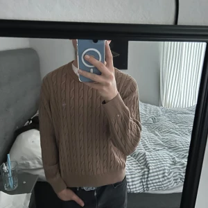 Beige kabelstickad tröja från GANT - Säljer en beige kabelstickad tröja från GANT med rund halsringning. Tröjan har ett diskret GANT-emblem broderat på bröstet.