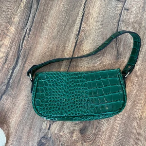 Grön axelväska i krokodilpräglat mönster - Säljer en snygg grön axelväska med krokodilpräglat mönster. Väskan har en kort axelrem och stängs med dragkedja. 