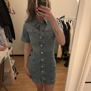 Jeansklänning med kort ärm och knappar - Säljer en snygg jeansklänning med korta ärmar och knappar hela vägen framtill. Klänningen har klassisk skjortkrage och två bröstfickor. Figurnära passform och kort längd, perfekt för en trendig look. Använd endast 1 gång så är i nyskick!!!☀️slutsåld från Zara
