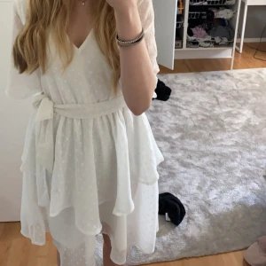 Vit omlottklänning med volang och knytband - Superfin vit omlottklänning från H&M med volangdetaljer och knytband i midjan. Klänningen har v-ringning, korta ärmar och ett diskret prickigt mönster i tyget. Perfekt för sommarens alla tillfällen!