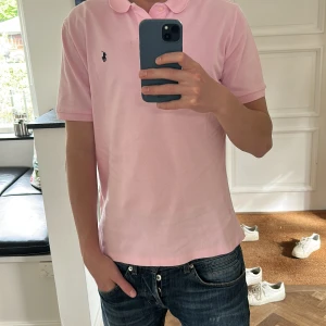 Ljusrosa pikétröja från Polo Ralph Lauren - Säljer en klassisk ljusrosa pikétröja från Polo Ralph Lauren med liten broderad logga på bröstet. Tröjan har korta ärmar, krage och knappar vid halsen. Perfekt för en stilren och avslappnad look.