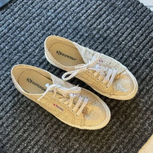 Glittriga sneakers från Superga - Säljer ett par snygga sneakers från Superga i glittrigt guld med vita snören och platt sula. Perfekta för dig som vill sticka ut lite extra! Klassisk modell med rund tå och Superga-logga på sidan.💗 använd två gånger 